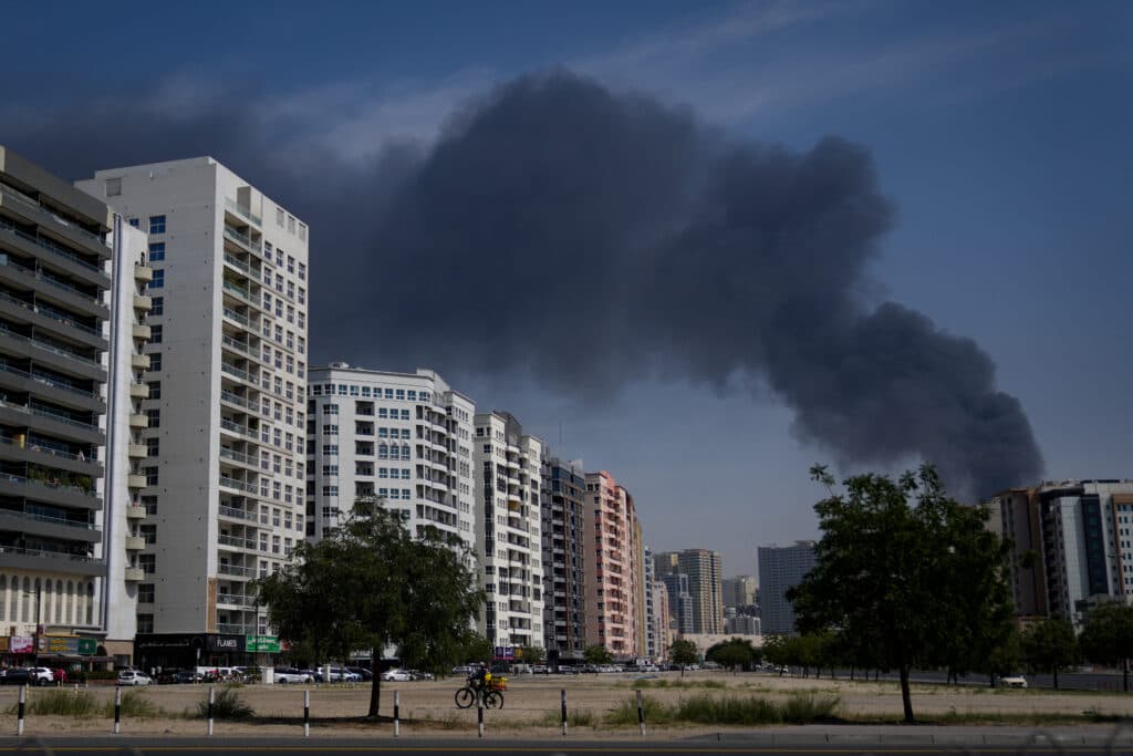 A large plume of black smoke rises into the sky above a cityscape featuring several multi-story residential buildings, under a clear blue sky.