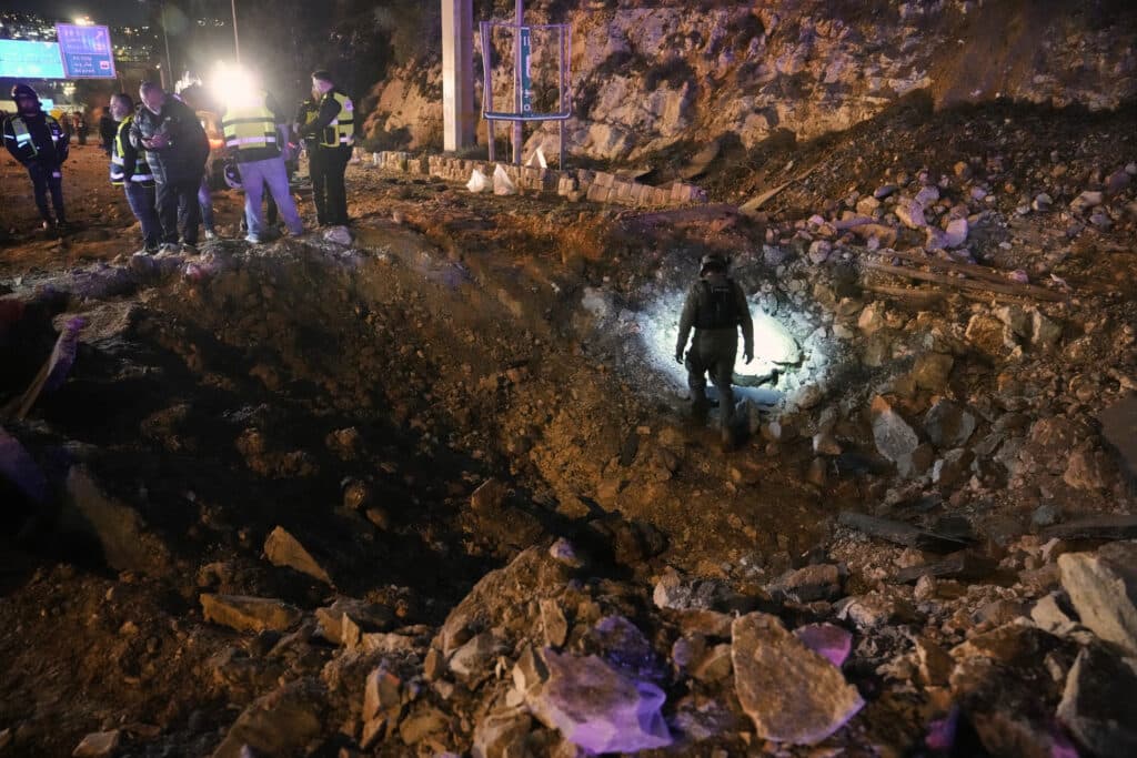 A group of people in safety vests stand beside a large crater at a construction site at night, while a person inspects the hole using a flashlight.