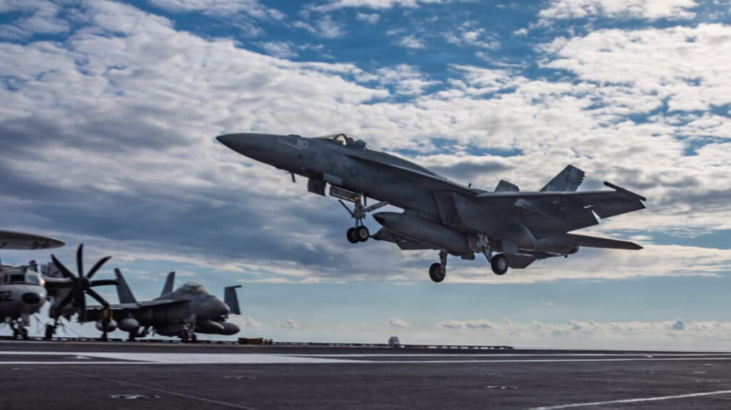 A fighter jet taking off from an aircraft carrier, with other planes parked on the deck under a partly cloudy sky.