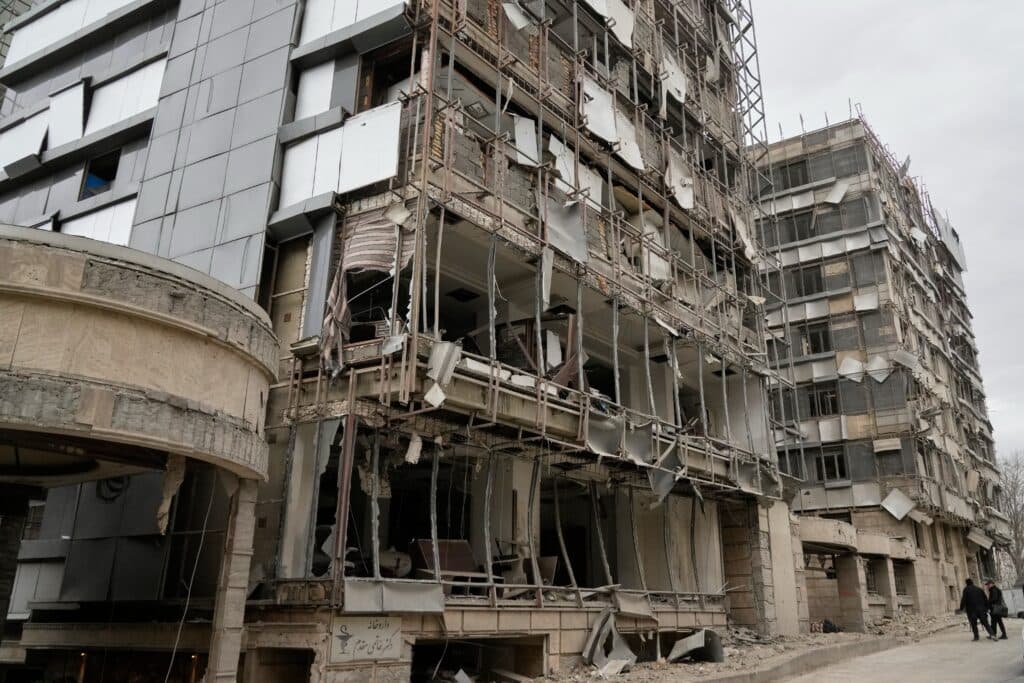 A damaged building with a partially collapsed facade and exposed interiors, showing extensive destruction, possibly from an explosion or natural disaster. Two people walk on the sidewalk nearby.