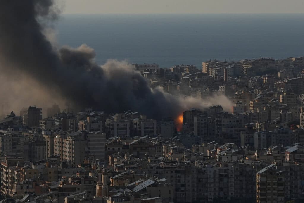 A cityscape with thick black smoke rising from a fire among densely packed buildings, set against a backdrop of the sea.
