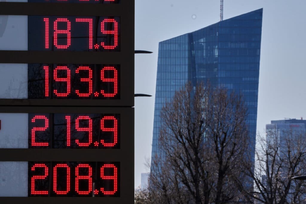 A digital display sign shows various fuel prices in bright red numbers, set against the backdrop of a modern glass skyscraper and leafless trees.