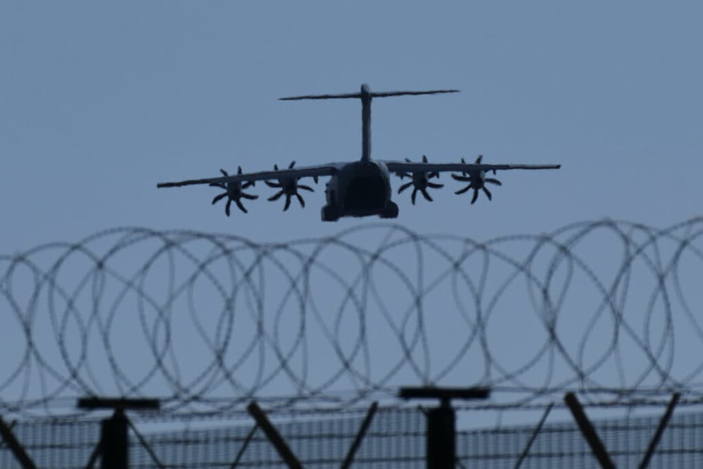 A military aircraft with four propeller engines approaching for landing over a barbed wire fence under a clear sky.