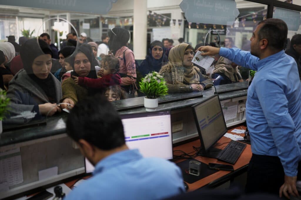 People wait in line at a busy service counter, with staff assisting them; a digital display and potted plants are visible on the counter.