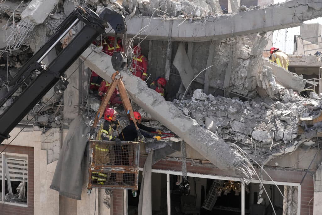 Rescue workers in red helmets and protective gear navigate the rubble of a collapsed building using a crane basket, amidst a scene of concrete debris and broken iron rods.