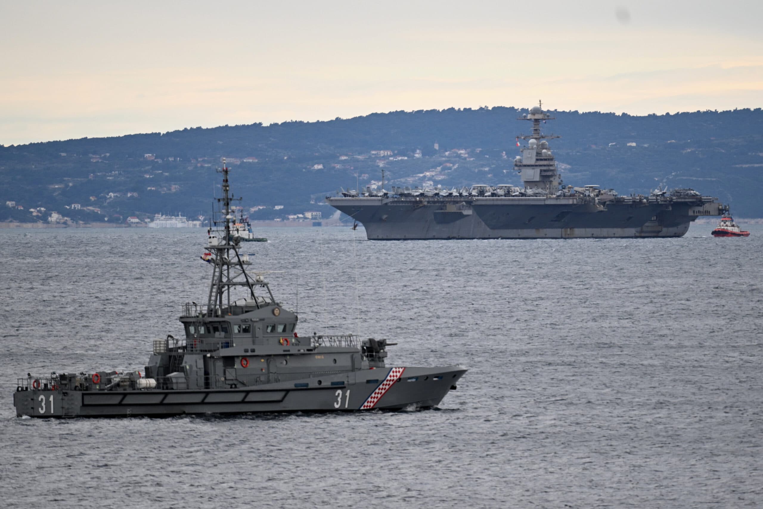 A naval ship with the number 31 visible on its side is sailing in the foreground, while a large aircraft carrier is seen in the background on open water, against a backdrop of distant hills and a cloudy sky.