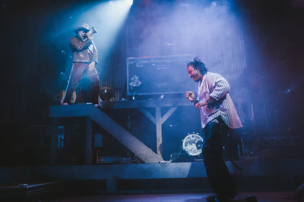 Two performers on stage: one standing on a platform with a megaphone and wearing a hat, and the other walking on the ground smiling, with stage lights and equipment in the background.