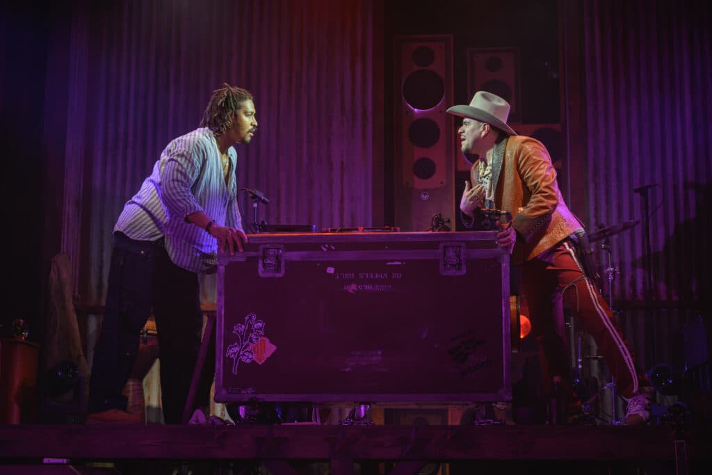 Two people engaged in a dramatic scene on stage; one wears a cowboy hat and an orange jacket, while the other wears a striped shirt. They are leaning over a large equipment case, with speakers in the background and a colorful lighting setup.