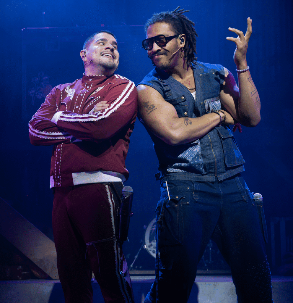 Two performers on stage, one wearing a red tracksuit and the other in a denim vest and jeans, both striking confident poses with crossed arms and microphones at their waists.