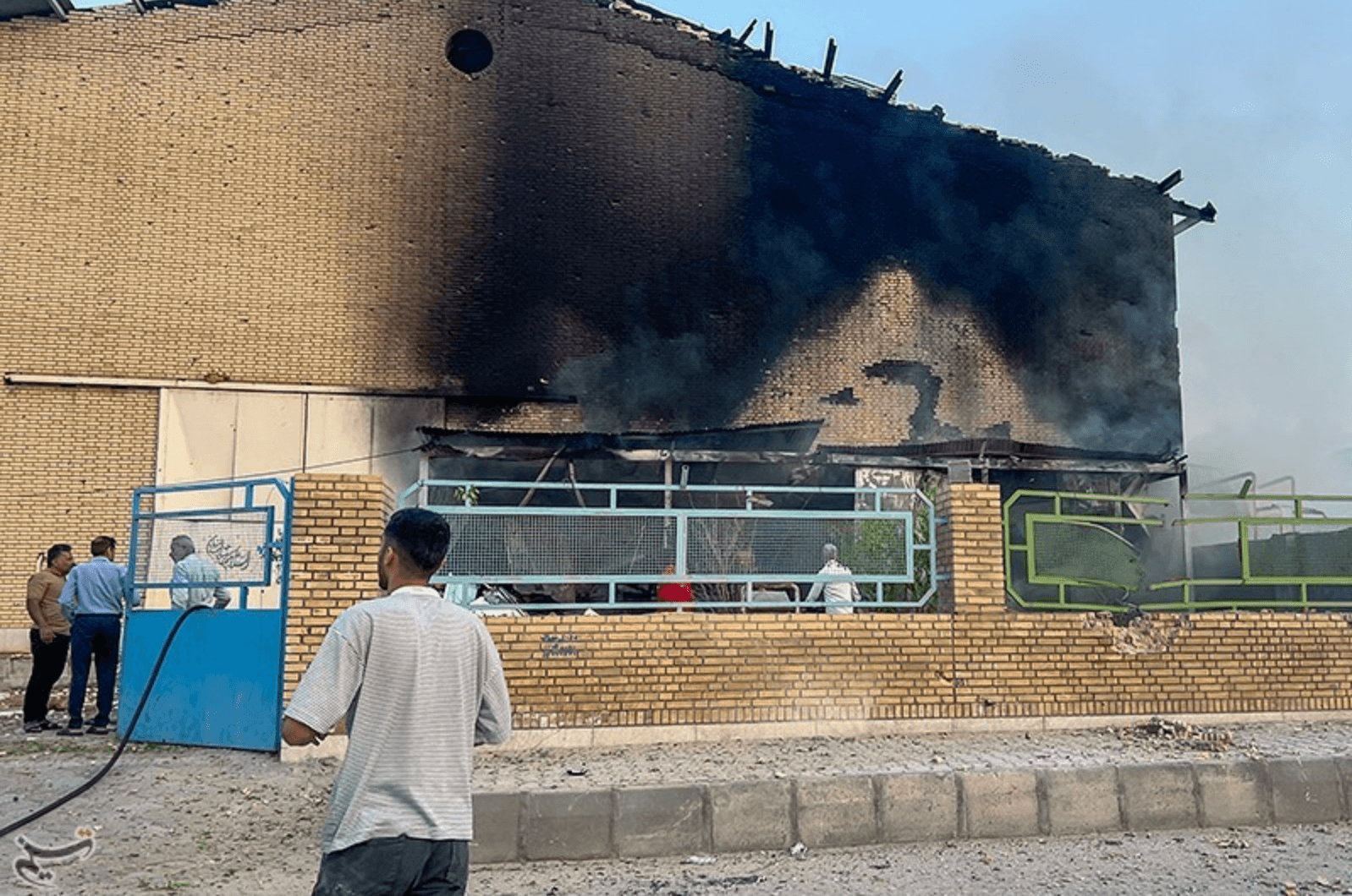 A brick building partially destroyed by fire, with a blackened wall and smoke still rising. Several people stand nearby, observing the scene, and a man in the foreground holds a hose, indicating efforts to extinguish the fire.