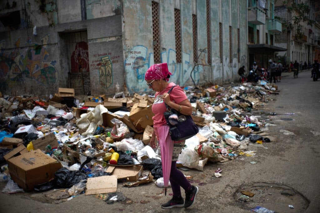 A woman in a pink shirt and purple leggings walks past a large pile of garbage on a city street, with graffiti-covered walls in the background.