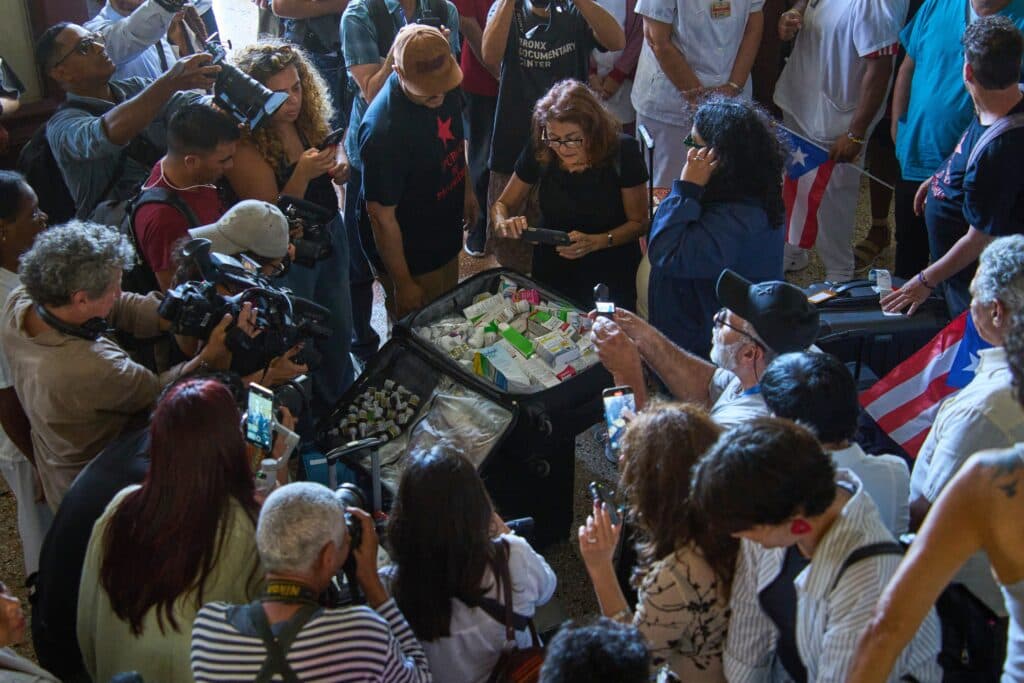 A group of people gathered around a suitcase filled with medical supplies, taking photos and videos. Some individuals are holding cameras and smartphones, while others hold flags. The setting appears to be indoors, with a diverse crowd showing interest and attention towards the contents of the suitcase.