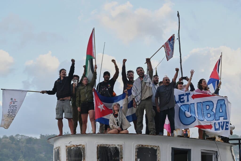 A diverse group of people standing on a boat, raising their fists and holding flags, including one reading "Let Cuba Live."