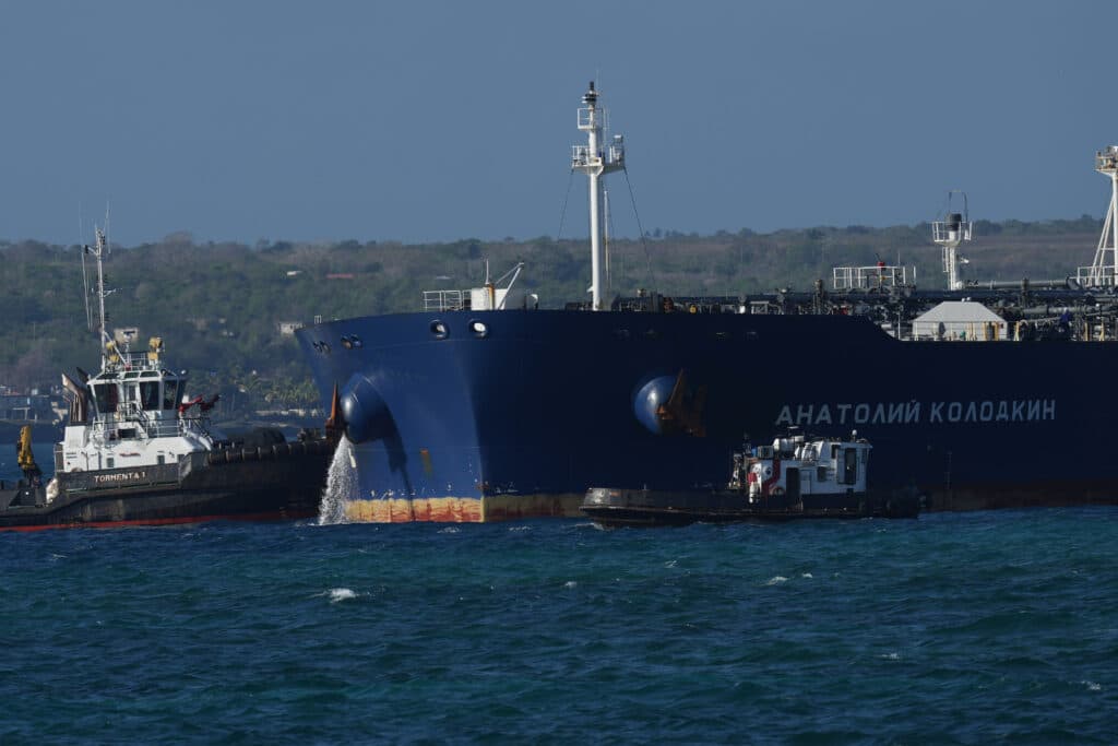 A large blue cargo ship with Cyrillic writing on its side, assisted by two smaller tugboats, navigating through a stretch of water with a distant shoreline in the background.