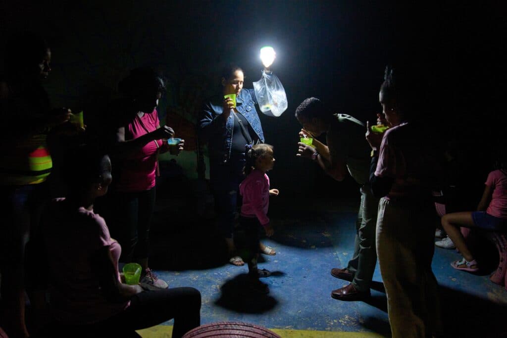 A group of people are illuminated by a bright bulb hanging in a dark room, holding green cups. A young child in a pink jacket is in the center, interacting with an adult bent over speaking to her. The setting appears intimate and communal, and the light casts dramatic shadows around the scene.