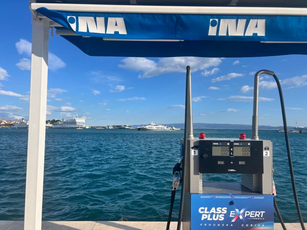 A fuel pump station by a body of water with a blue awning marked 'INA'. In the background, several large ships are visible in the harbor under a blue sky with scattered clouds.
