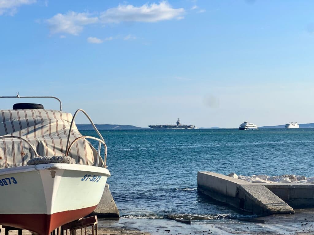 A boat with striped covering is docked on the shore with a view of the sea, where larger ships are visible in the distance under a clear blue sky.