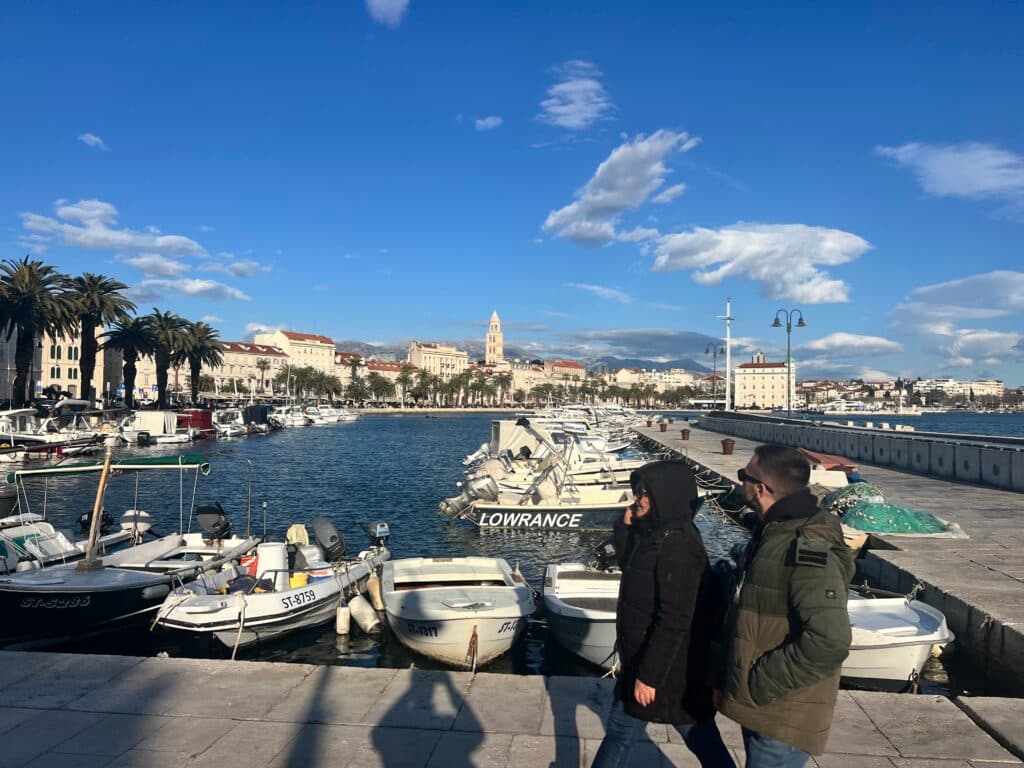 A coastal promenade lined with palm trees and boats docked along the water. A clear blue sky with scattered clouds is overhead, and a couple wearing jackets walks along the pier. Historic buildings and a clock tower are visible in the background.
