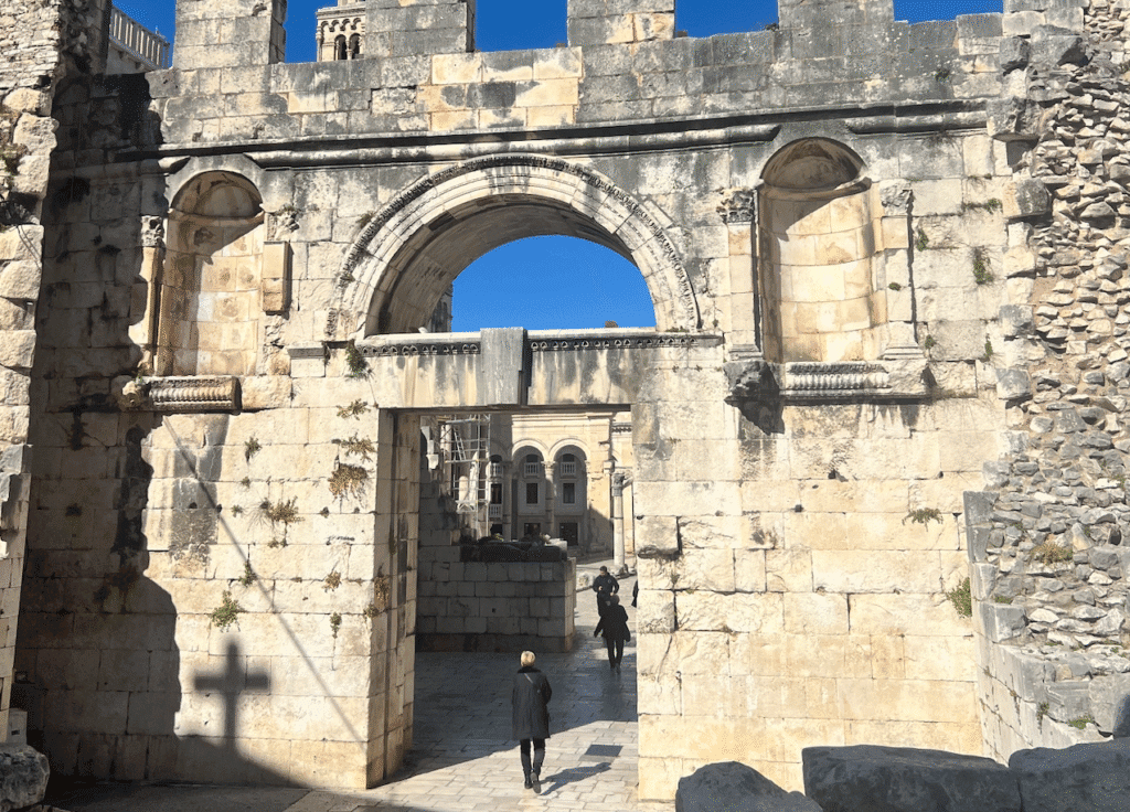 An ancient stone archway with historical architectural details, featuring plants growing in crevices. A person is walking through the archway, and a shadow of a cross is cast on the left side of the structure. Behind the arch, a modern building is visible under a clear blue sky.