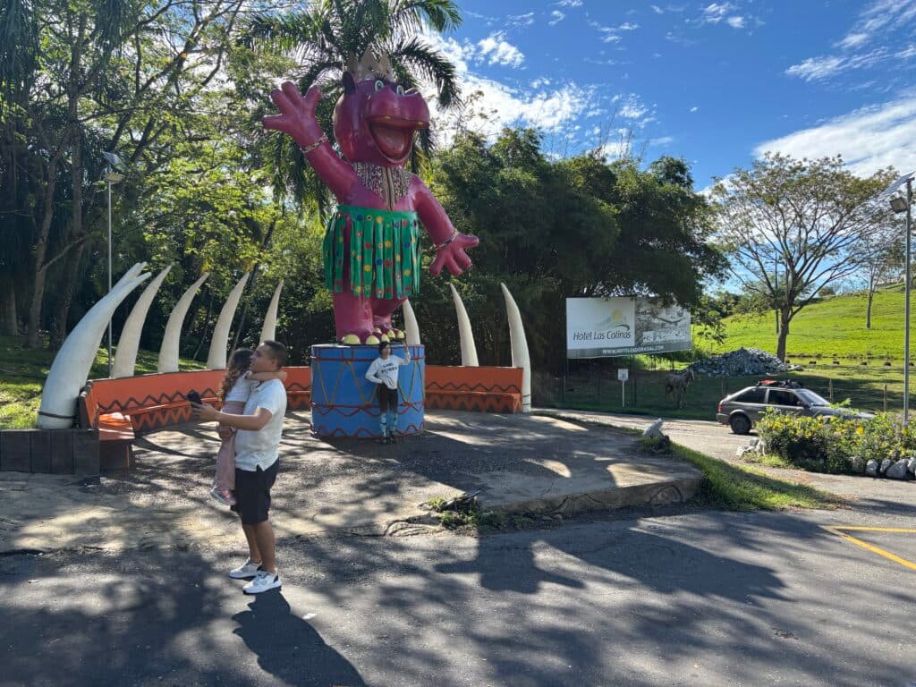 A person stands near a large, colorful dinosaur statue in a tropical outdoor park.