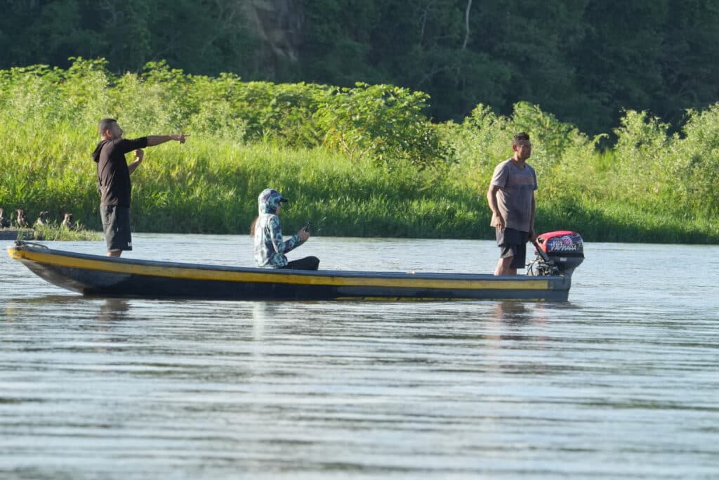 Three people in a small motorboat on a river, with one standing and pointing while another operates the motor.
