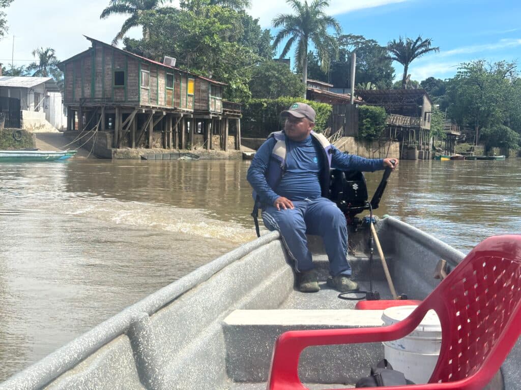 A man operating a motorboat on a river with stilt houses and trees in the background.