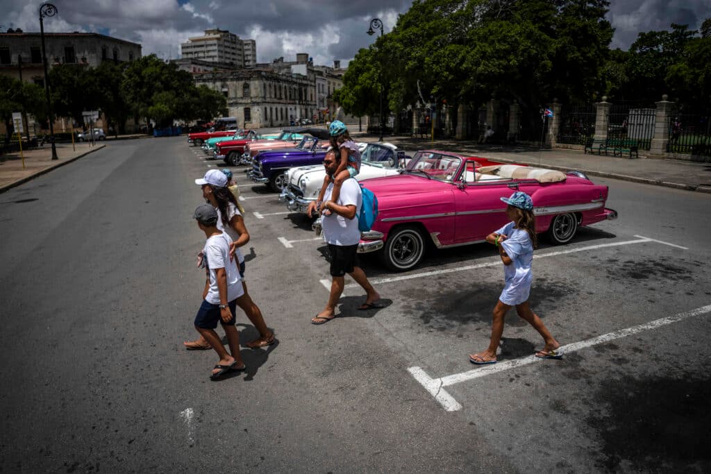 A family crosses a street in front of a lineup of colorful classic cars parked in an urban setting under a partly cloudy sky.