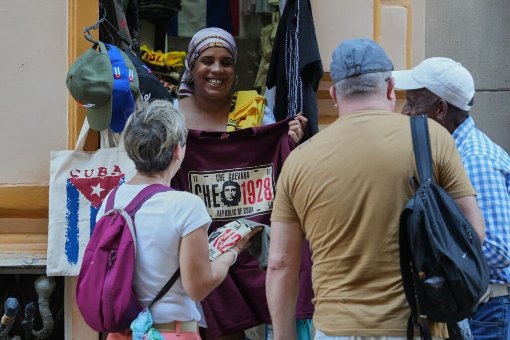 A street vendor shows a Che Guevara shirt to three tourists near bags with a Cuban flag design.