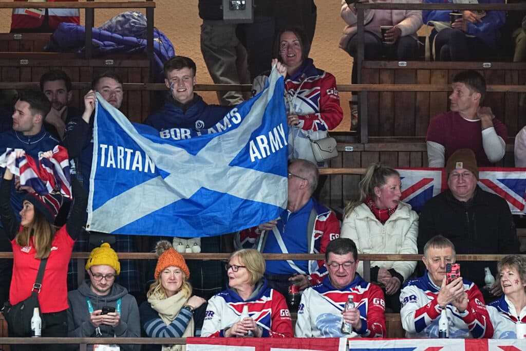 A group of people wearing hockey jerseys and holding a Scottish flag with "Tartan Army" written on it, in a wooden arena setting.