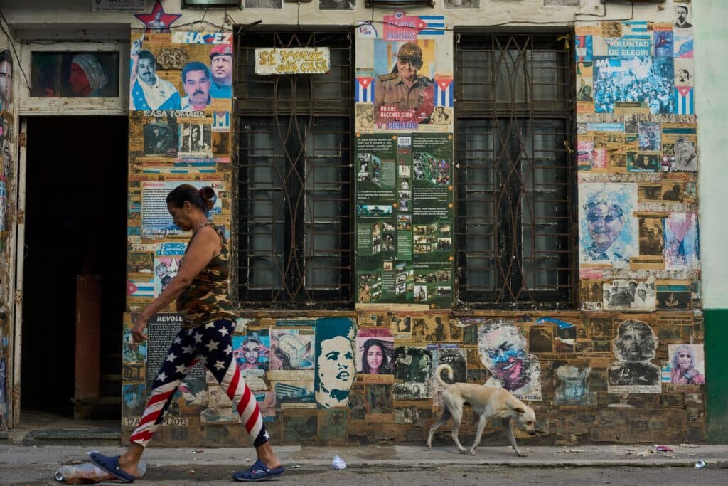 A woman walks past a wall covered in colorful posters and images, accompanied by a small dog on the street.