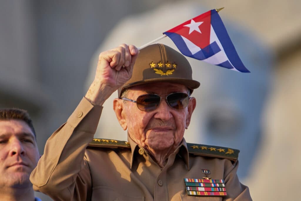 An elderly man in a military uniform salutes while holding a small Cuban flag.