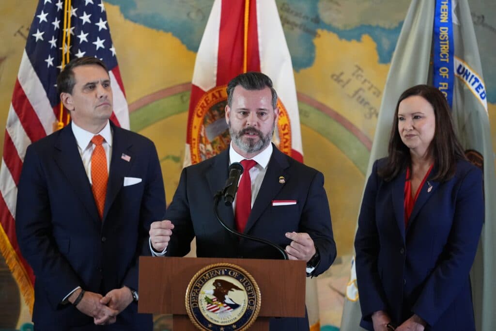 Three officials stand in front of flags and a podium with a Department of Justice seal, against a backdrop of a colorful mural.