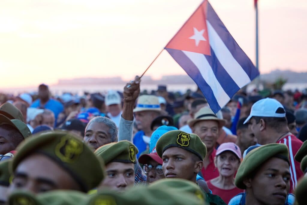 A crowd of people, some in military-style hats, with one person holding a Cuban flag.
