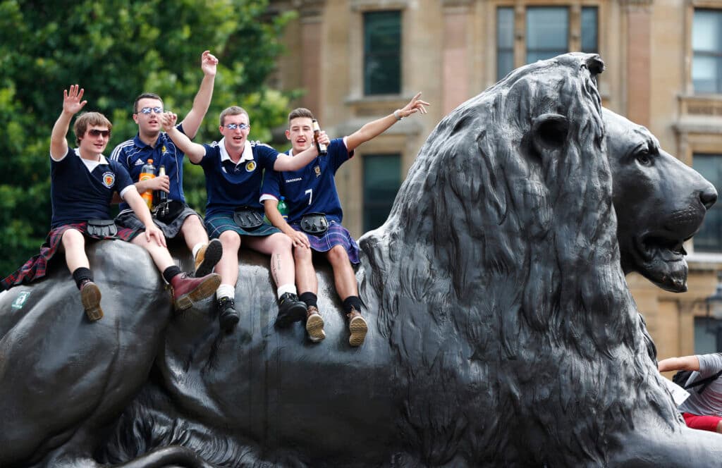 Four men wearing kilts and Scotland shirts sit on a large lion statue, waving and smiling.
