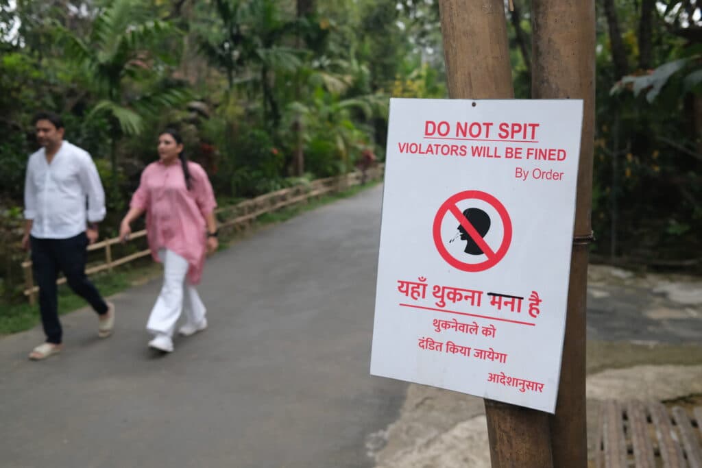 Sign warning against spitting with a fine, featuring text in English and another language, near a path in a park.