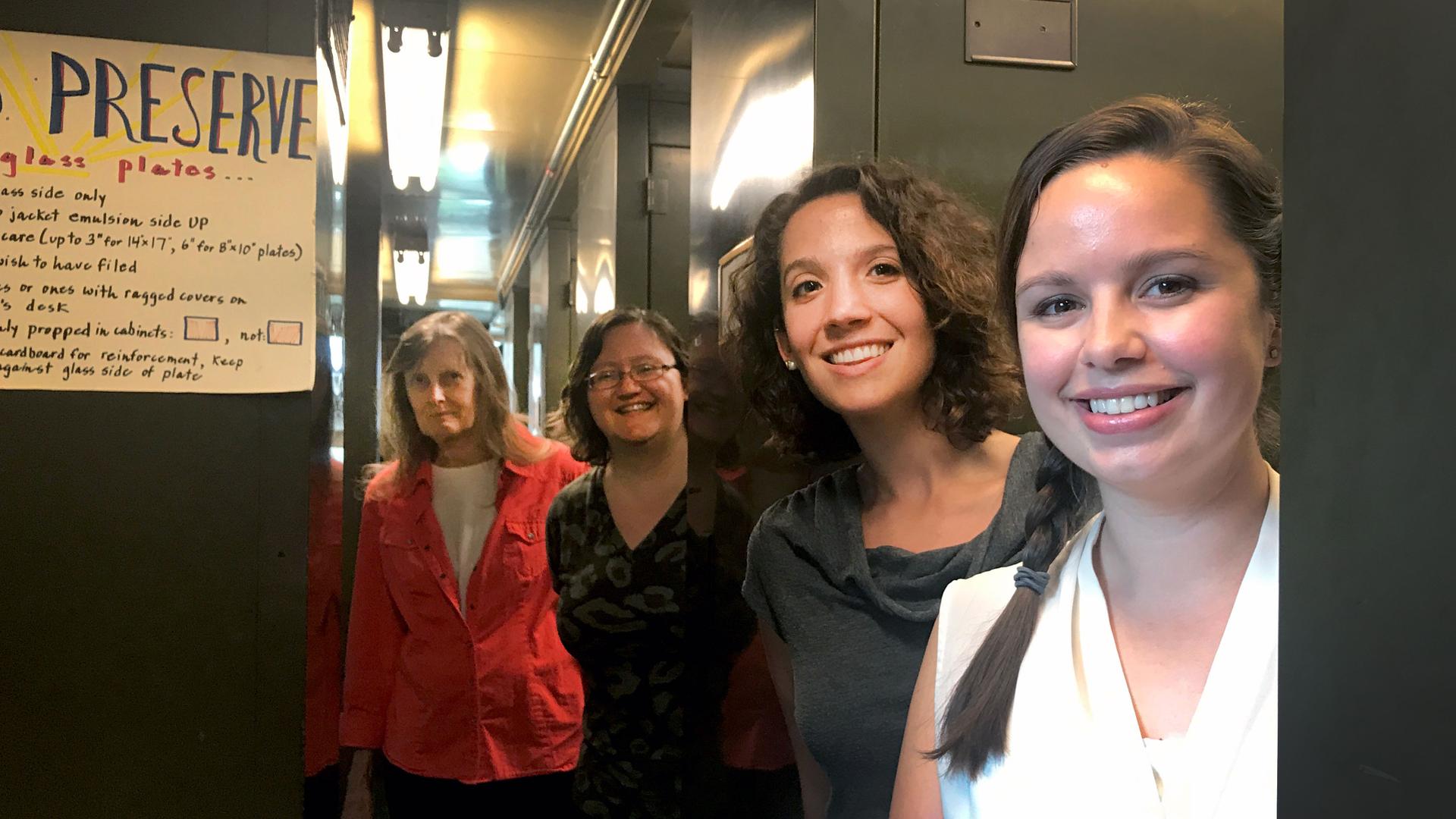A group of four women pose in the Harvard plate stacks, surrounded by tall metal cabinets. A paper sign to their left says 
