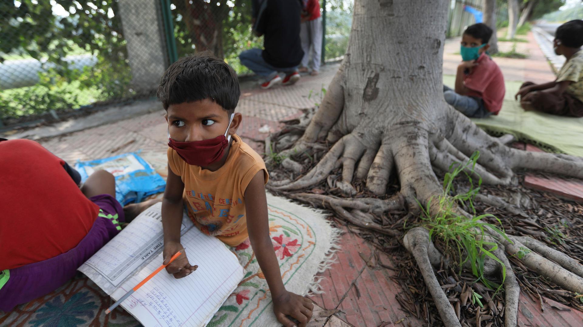 A young boy is shown wearing a red mask and holding a pencil while sitting on a mat next to a tree outside for school.