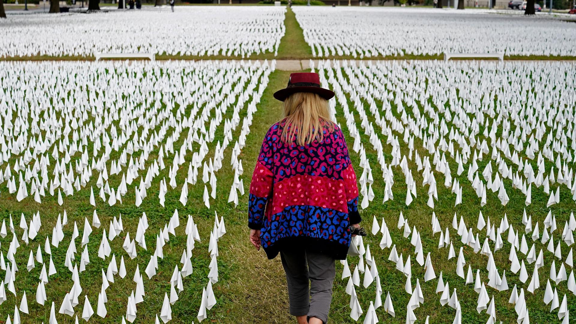 A woman wearing a colorful sweater and brimmed hat is shown from behind walking in a narrow path on a lawn filled with small white flags.