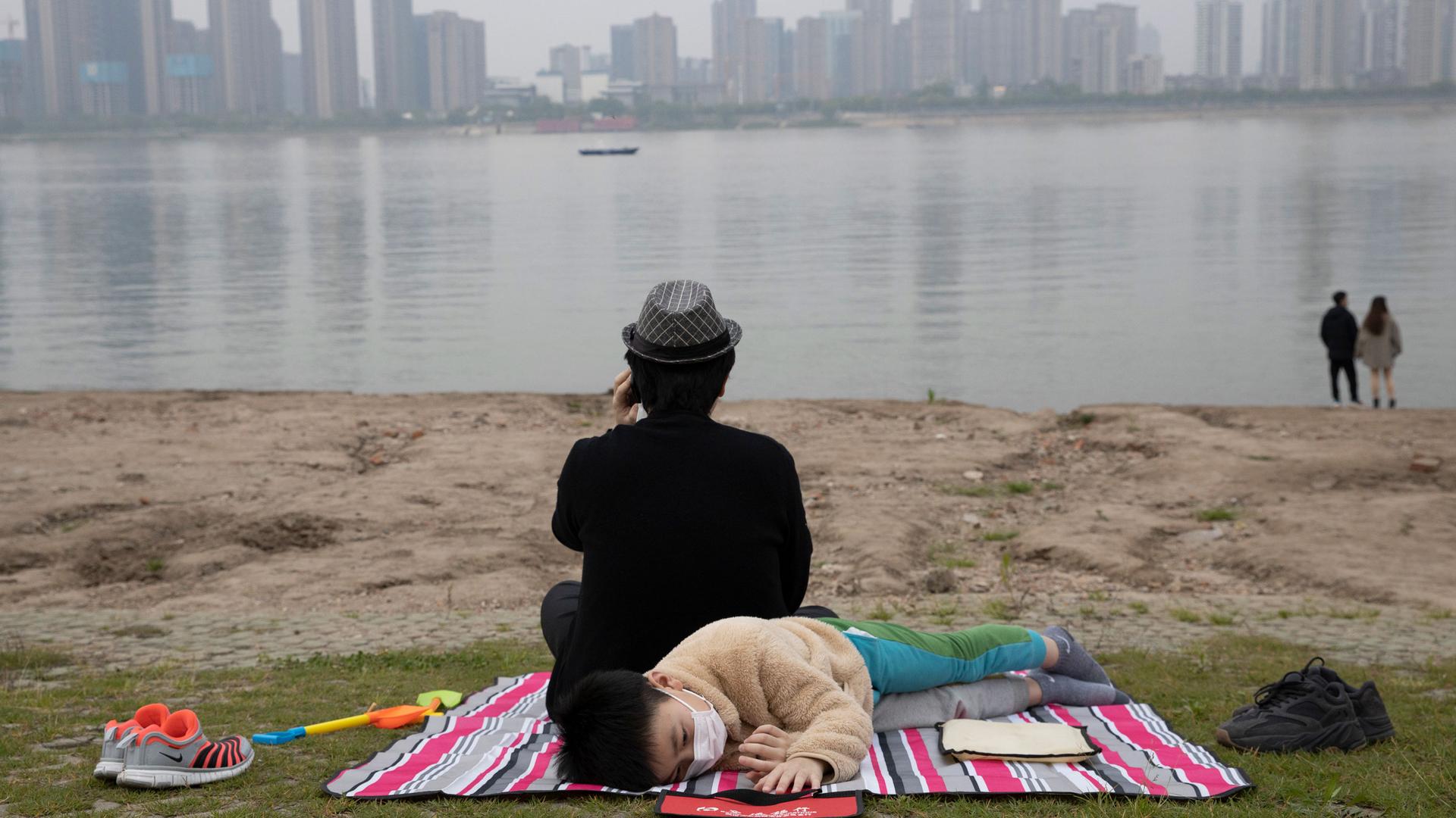 A young child is shown laying on a striped blanket and wearing a mask with a man sitting and facing the opposite direction looks on at the Yangtze River.