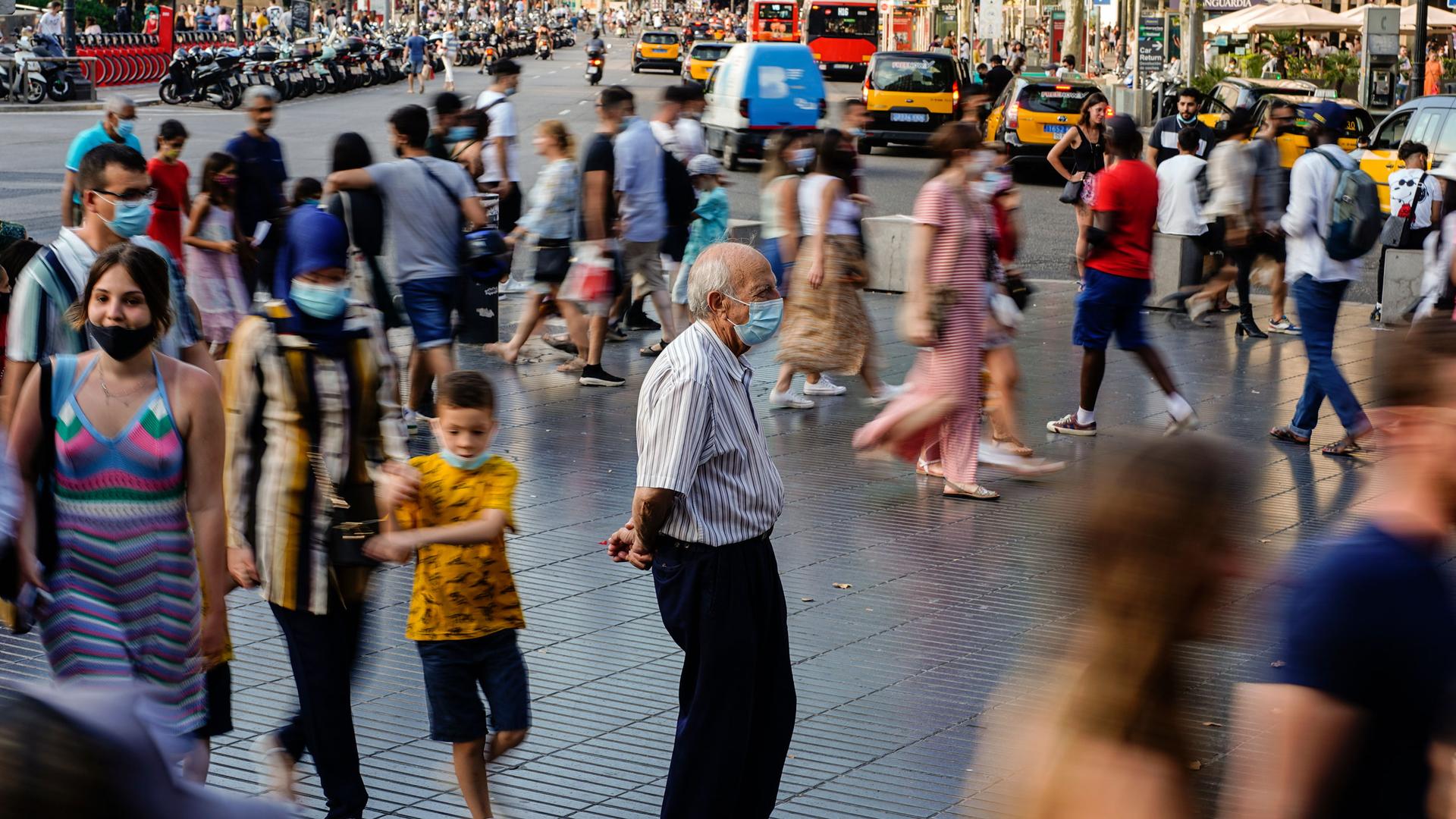 An older man is shown standing still in a crowded sideway area with people walking past him in blurred motion.