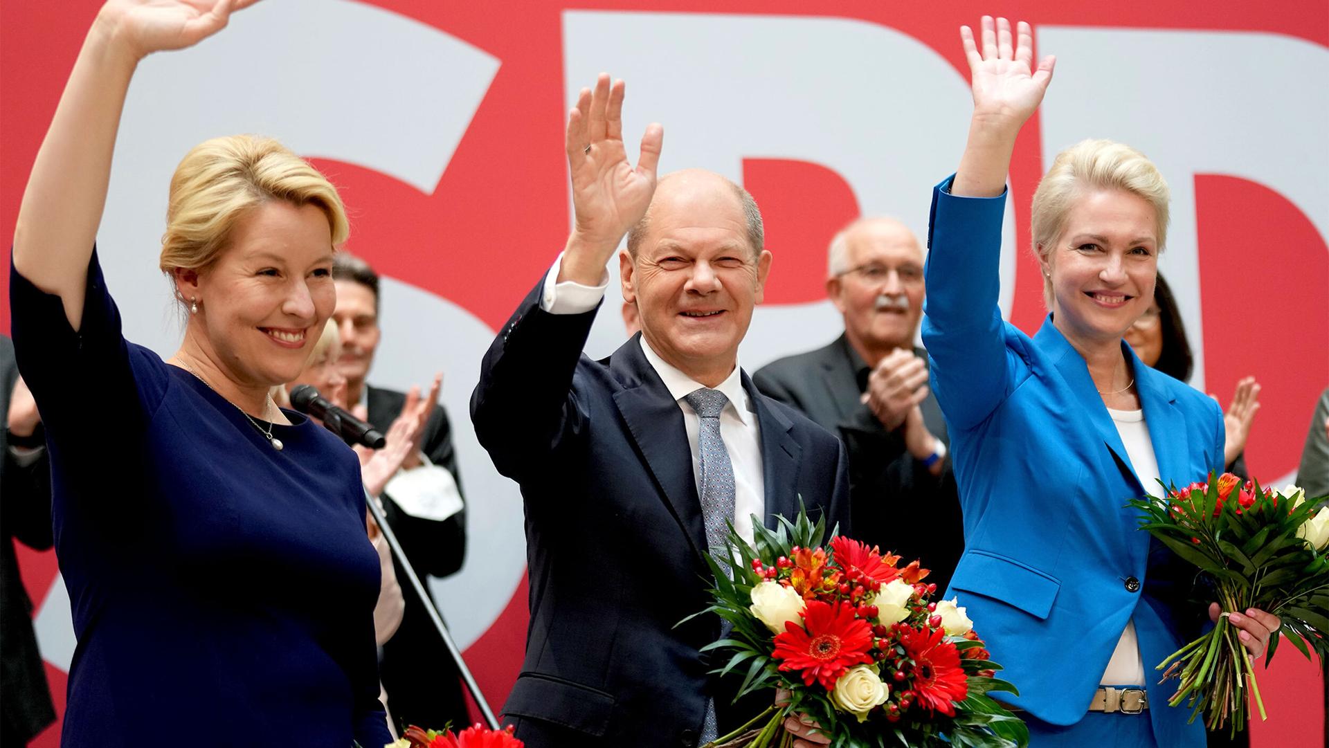 Olaf Scholz is shown wearing a dark suit and standing inbetween two other top German politicians with all three waving their right hands.